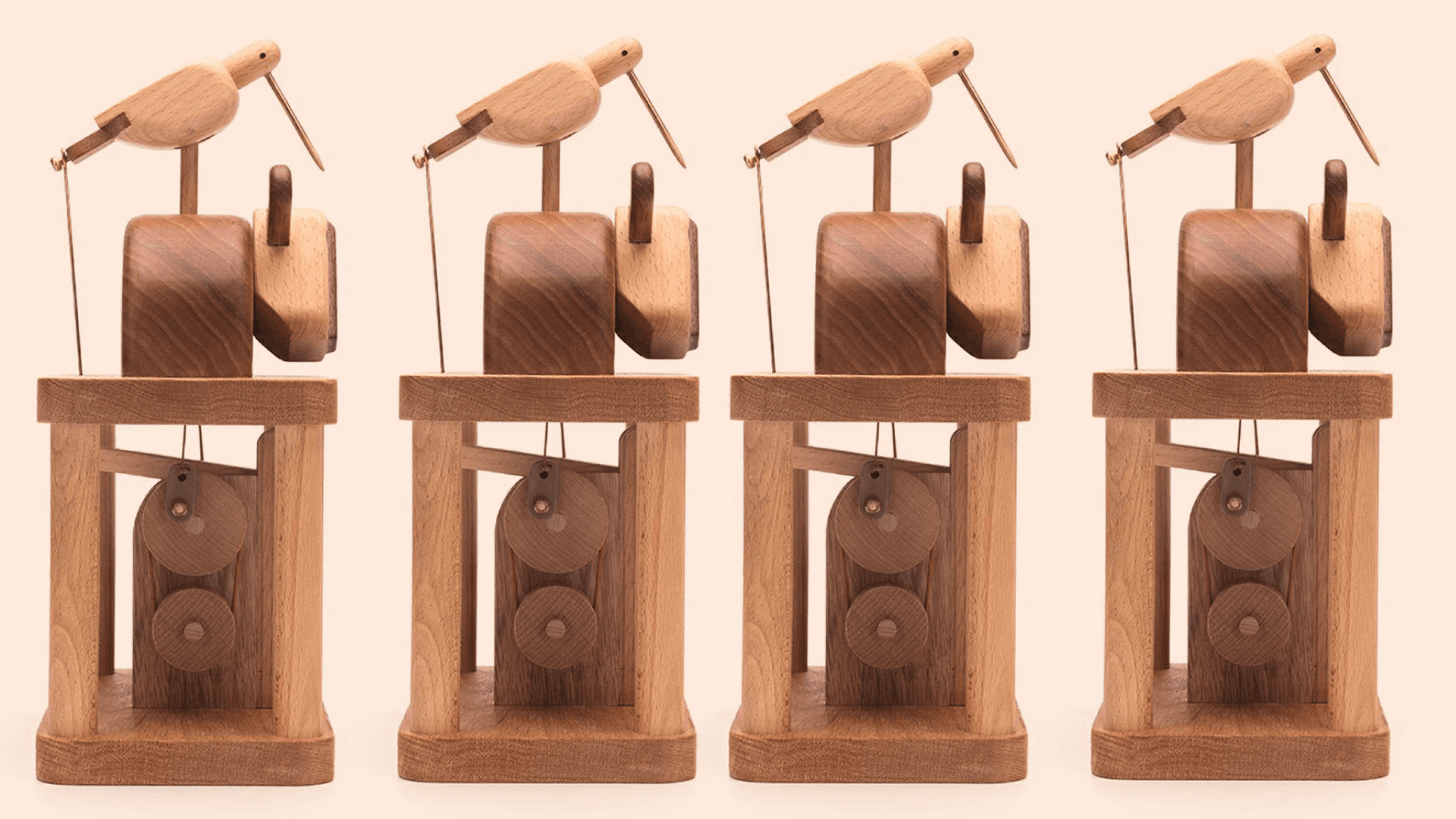 Fotografía. Sobre un fondo beige se presentan cuatro autómatas idénticos, hechos principalmente de madera. Cada uno tiene una estructura de base rectangular, con dos poleas en la parte inferior. Encima de la estructura, hay una figura de un ave minimalista tallada en madera. El ave se encuentra con el pico apuntando hacia abajo, sostenida por dos cuerdas que permiten el movimiento al girar una manivela.