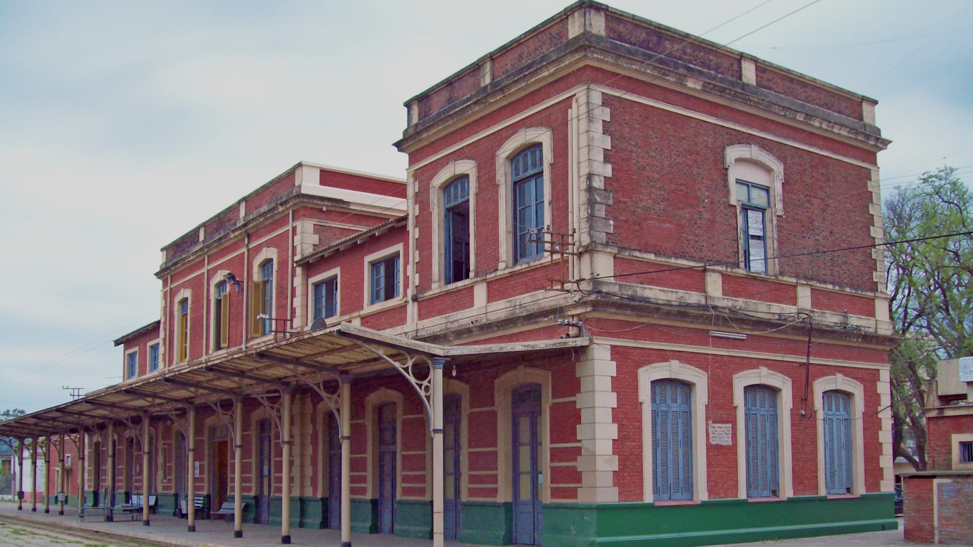 Ilustración. Sobre un fondo de cielo con nubes se observa una fotografía urbana con edificios de ladrillo y una antigua estación con techo metálico y rieles de tren en primer plano.