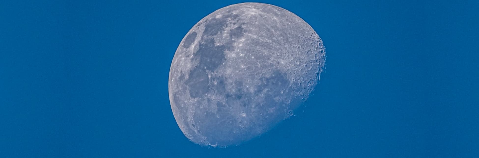 Foto. Sobre un fondo azul claro aparece muy de cerca la luna con sus cráteres y mares.