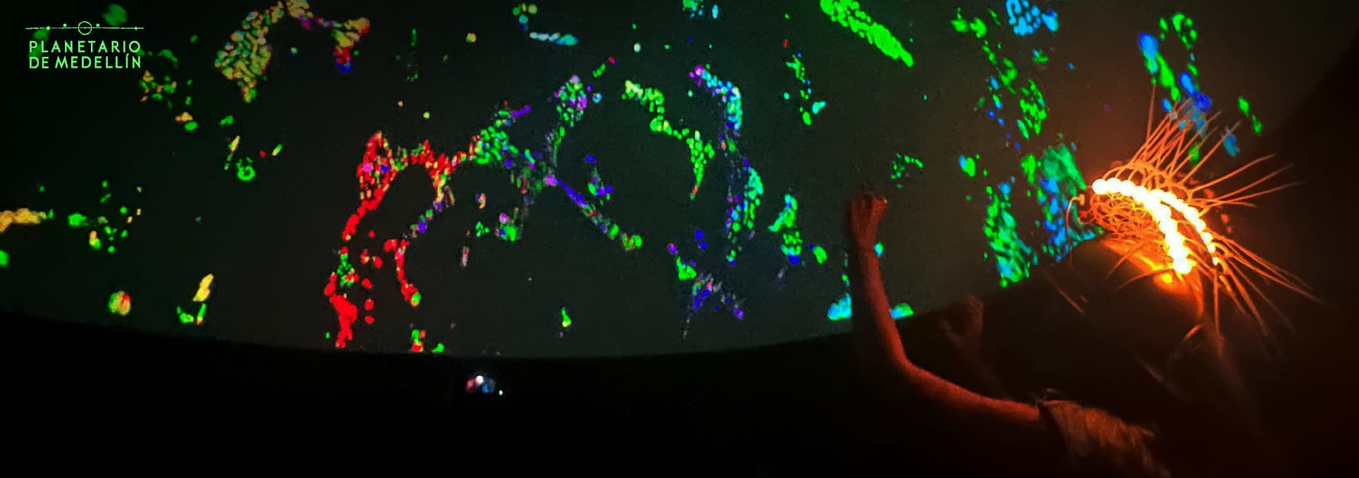 Fotografía. En una sala oscura con proyección en cúpula, se observan patrones luminosos en colores verdes, rojos, azules y violetas. En el borde inferior derecho, una persona usa un casco decorado con luces anaranjadas y levanta el brazo hacia la proyección.