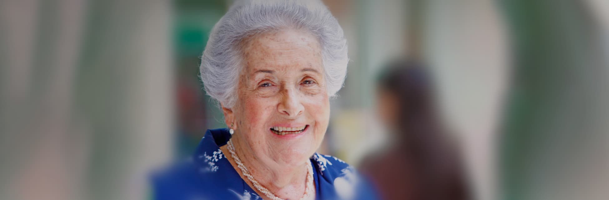 Foto horizontal. En el centro de la imagen un primer plano del rostro de la doctora Ángela Restrepo Moreno, está de medio perfil. Tiene el cabello blanco y corto. Cejas cafés delgadas, ojos pequeños. Está sonriendo.