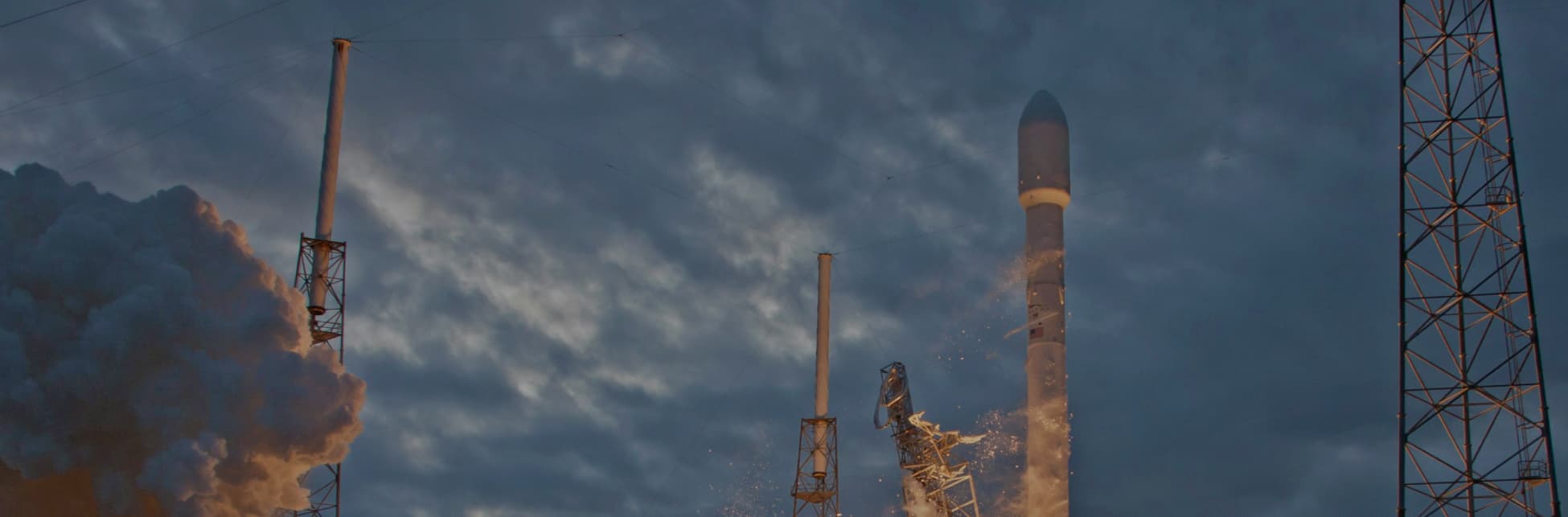 Fotografía. En el centro, un cohete en proceso de despegue, con fuego y humo saliendo de su base. El cielo de fondo muestra una mezcla de nubes y luz tenue.