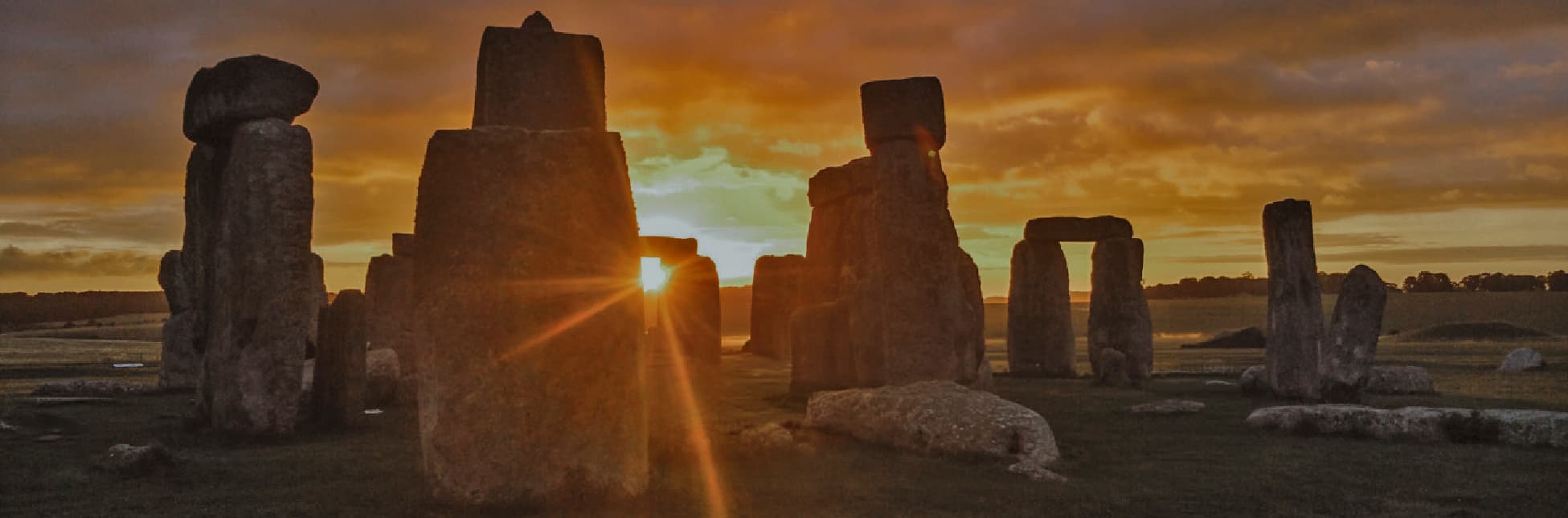 Fotografía. Un conjunto de grandes piedras verticales se recorta frente a un atardecer anaranjado; el sol se asoma entre los bloques y proyecta un destello hacia la cámara