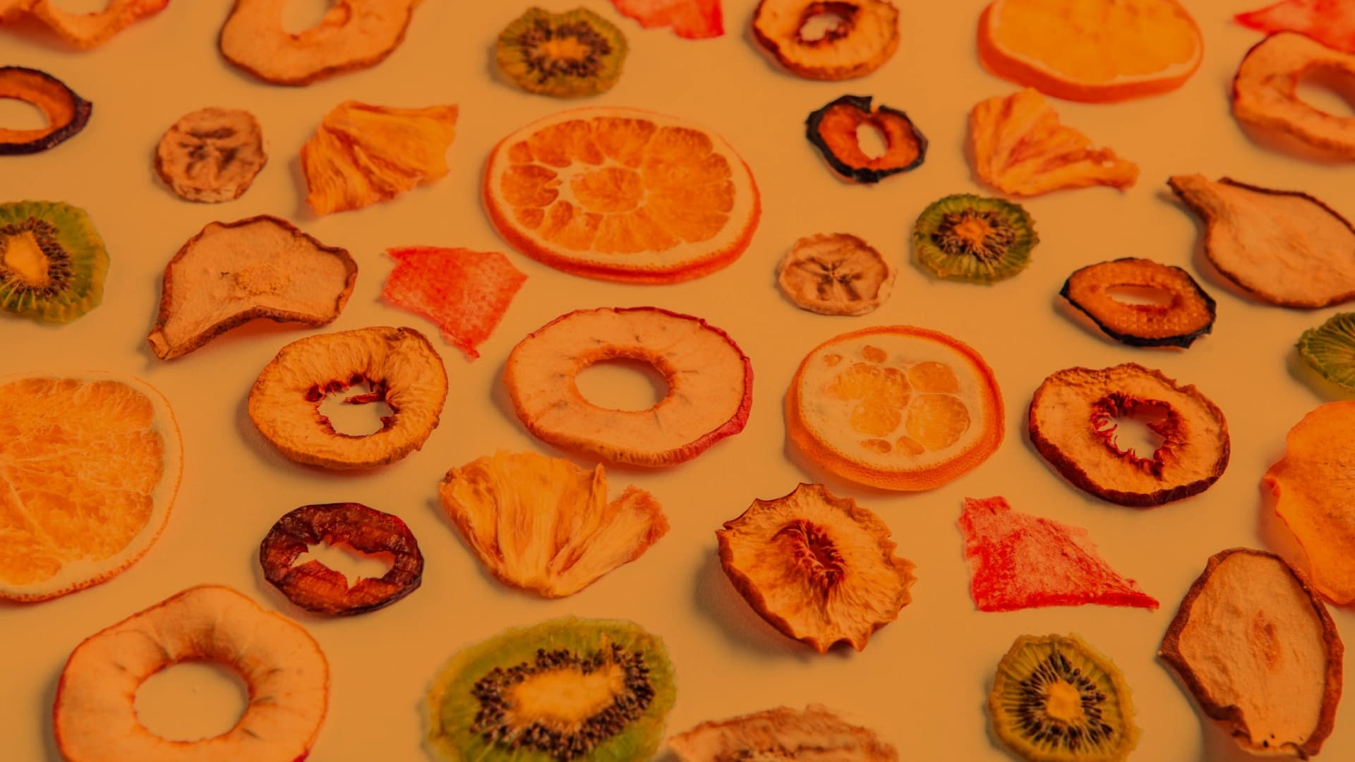 Fotografía. Sobre un fondo claro se disponen múltiples rodajas y fragmentos de frutas deshidratadas distribuidas de manera uniforme. Se observan círculos completos y anillos con hueco en el centro, así como piezas irregulares. En la zona central hay una rodaja de naranja con sus gajos visibles, rodeada de otras frutas como kiwi en cortes circulares con semillas, manzana en anillos con borde más oscuro, y rodajas más pequeñas con texturas arrugadas. También aparecen fragmentos triangulares y láminas delgadas de fruta, algunas con bordes ondulados. Las piezas varían en tamaño y forma, creando un patrón repetido a lo largo de toda la superficie.