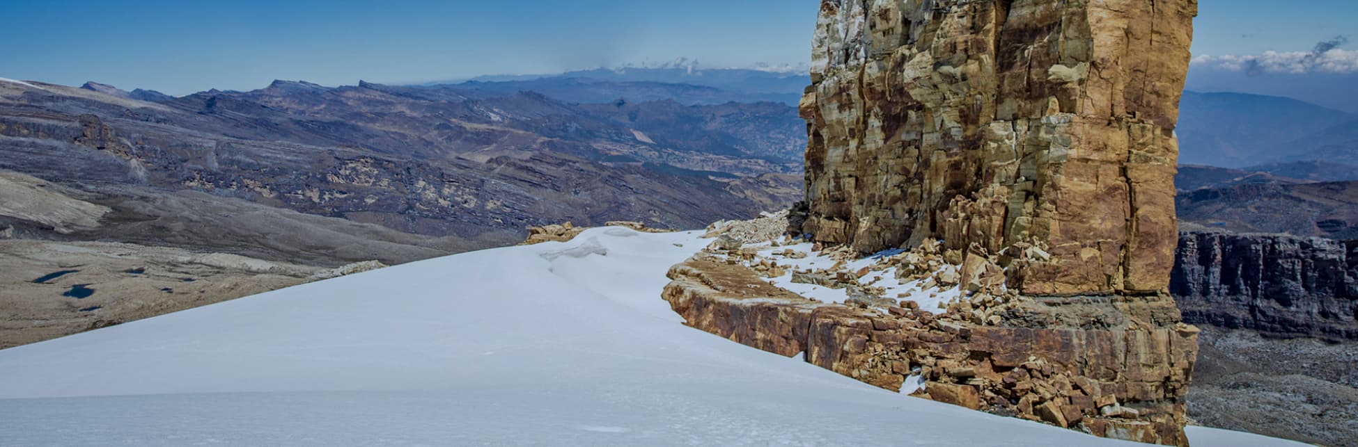 Foto. En la parte inferior de la imagen sobre un suelo cubierto de nieve aparece una formación rocosa de forma rectangular. Al fondo un valle desértico.
