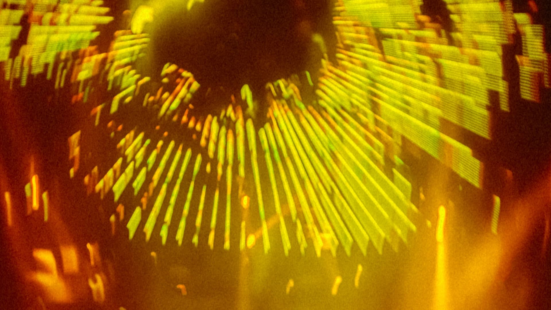 Fotografía. Vista abstracta con apariencia de proyección luminosa: desde una zona oscura central se expanden líneas radiales y fragmentos alargados, con destellos y formas desenfocadas alrededor que sugieren movimiento, reflejos y un efecto óptico envolvente.