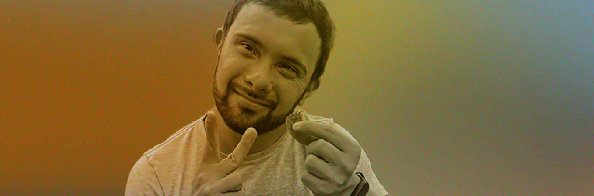 Ilustración. Sobre un fondo con degradado de varios tonos aparece, en la parte inferior derecha, la fotografía de un hombre joven con barba corta y cabello corto que sonríe mientras sostiene un pequeño objeto entre los dedos de una mano y señala hacia él con la otra.