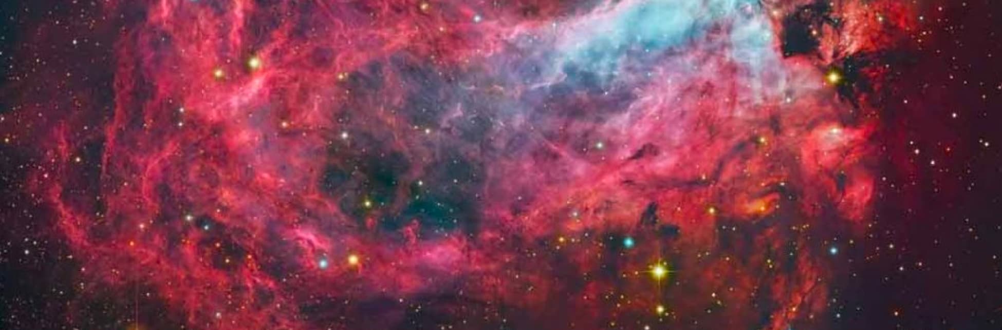 Foto. Sobre un fondo estrellado aparece una nebulosa, manchas dispersas en el cielo en tonos rosados y azules sobre las cuales se ven puntos de distintos colores unos más grandes que otros.