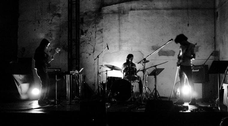 Fotografía. Sobre un escenario una banda de rock interpreta su música. Está conformada por un bajista, un guitarrista y una baterista.