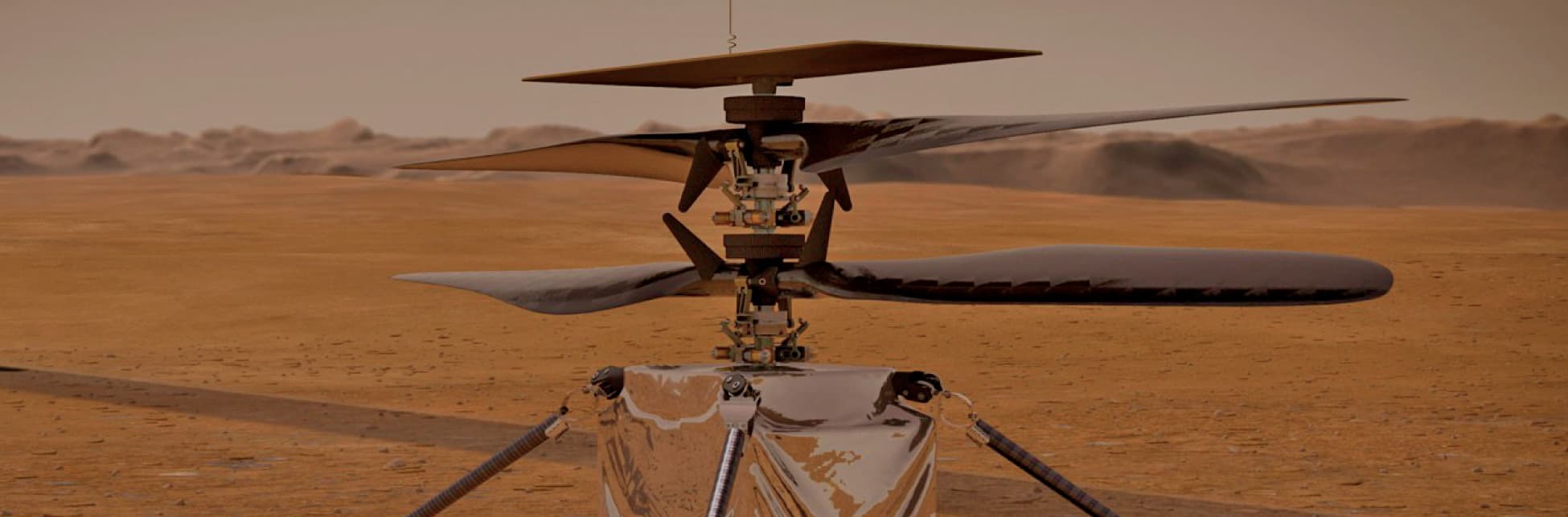 Fotografía. El fondo muestra un escenario desértico con montañas de arena. La imagen está centrada en el dron Ingenuity, que tiene cuatro aspas y un circuito en el medio de las mismas. En la parte inferior, hay una caja sostenida por cuatro soportes sobre la tierra.