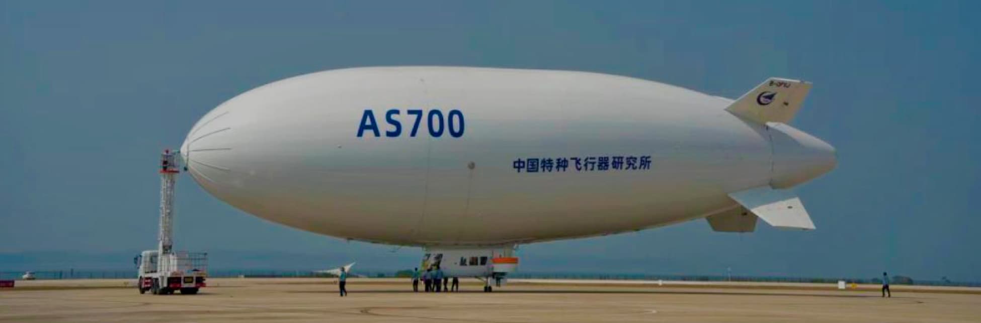 Fotografía. Un dirigible con forma cilíndrica y alargada de color blanco, marcado con las letras y números AS700, en la zona central de la aeronave varias personas abordan su cabina.