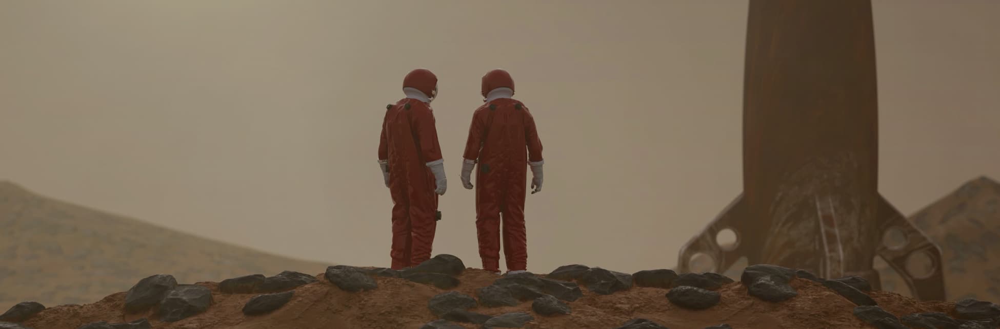 Foto. En la parte inferior de la imagen sobre una superficie rocosa y arenosa, dos personas de cuerpo entero, de espaldas, con traje de astronauta rojo. Frente a ellos un cohete.