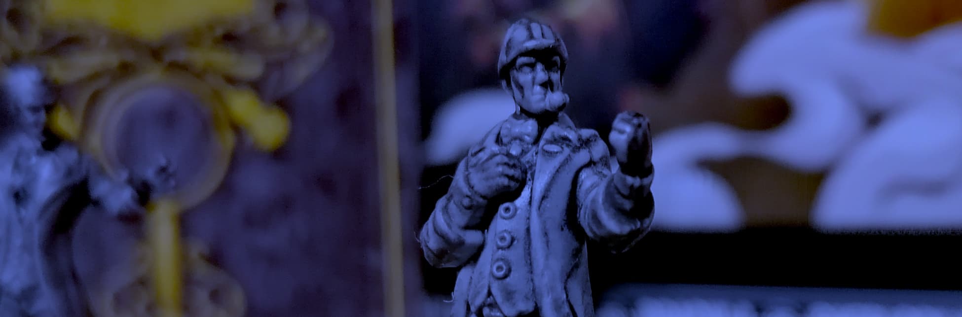Fotografía de una miniatura de un detective con sombrero y pipa, ubicada sobre una base circular. En el fondo, hay otras miniaturas y una ilustración de un tablero de juego con tonos oscuros y detalles dorados.