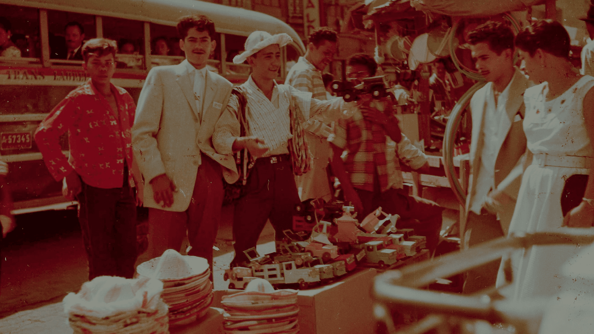 Fotografía. Un grupo de personas en un mercado popular. A la izquierda de la imagen hay cinco hombres, en el medio hay cajas con distintos productos como sombreros y carros de juguete. A la derecha hay un hombre de cachaco y una mujer de vestido que observan los productos.