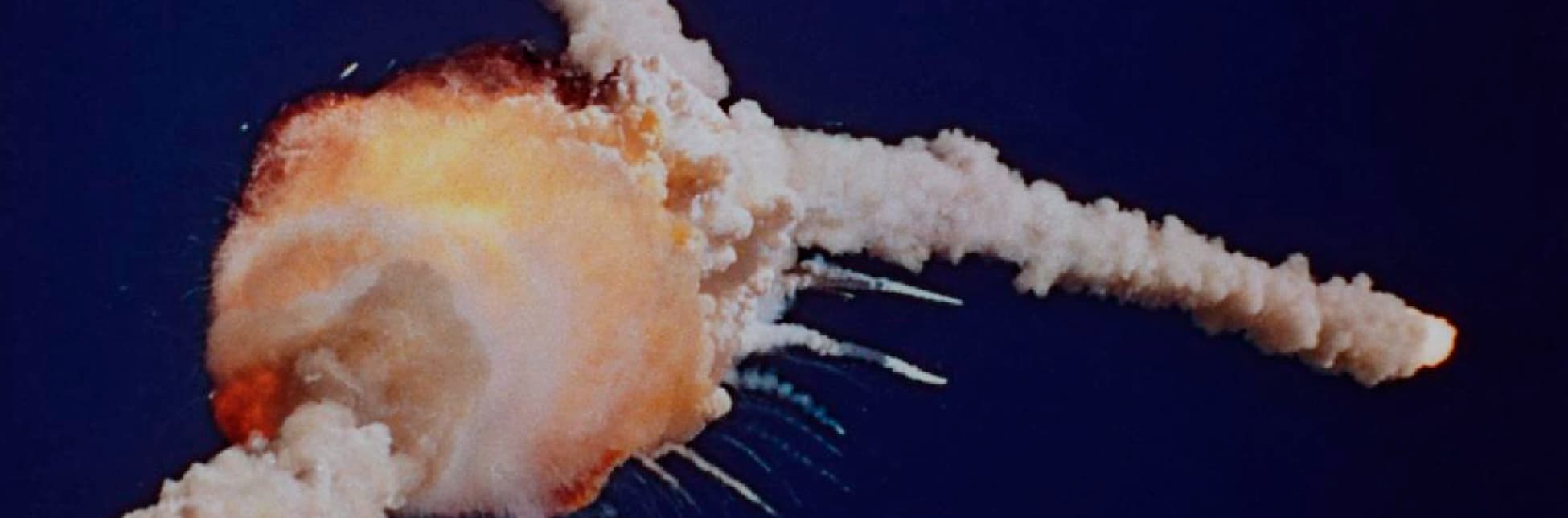 Fotografía. En el centro se observa una gran nube de humo blanco con tonos anaranjados y amarillos que se expande en distintas direcciones. La forma principal tiene apariencia de una explosión, con una esfera luminosa en el punto de origen y varias estelas de humo que se extienden hacia la derecha sobre un fondo azul oscuro. La escena transmite un momento de ruptura y dispersión en el aire.