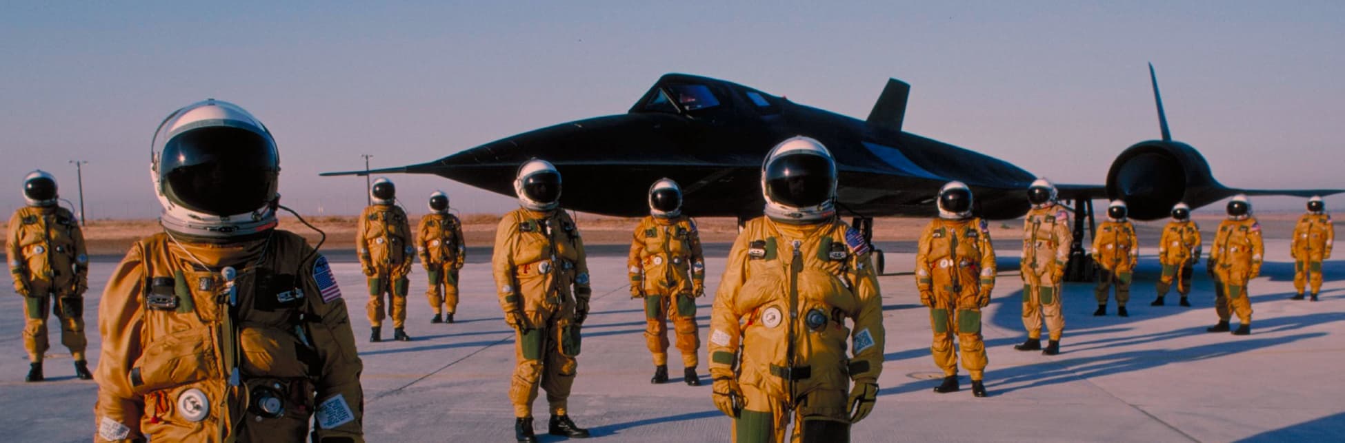 Foto. Varias personas con traje de astronauta y casco de pie, al fondo una nave color negro.