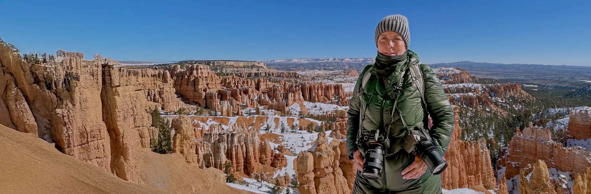 Fotografía. En el centro de la imagen una mujer con ropa de invierno con gorro y chaqueta, de su cuello cuelgan dos cámaras fotográficas. Al fondo varias formaciones rocosas cubiertas de hielo.