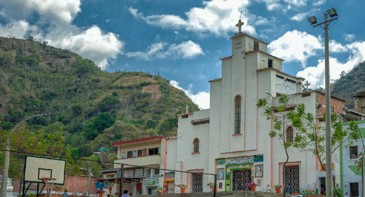 Foto. En la parte derecha el frontis de una iglesia. A la derecha una casa y al fondo una montañan. En la parte superior el cielo azul tiene algunas nubes.